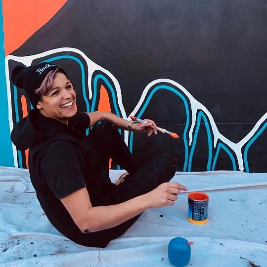 Artist Jasmine Sarin painting the NCIE mural, Jasmine sits on ground with big smile on face she is wearing a beanie and has a paintbrush in her hand, in front of her is the mural she is painting it features black, white, blue and orange lines